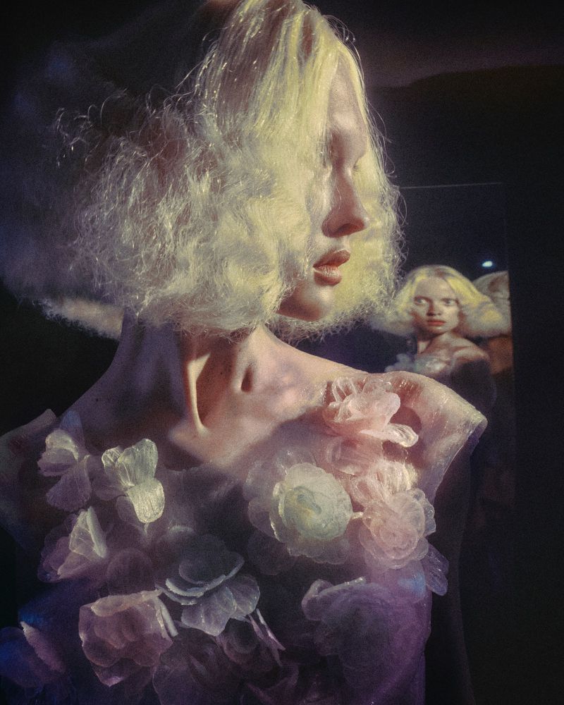 A woman with blonde hair and floral attire poses indoors with a blurred reflection in the background, showcasing an artistic and ethereal quality.