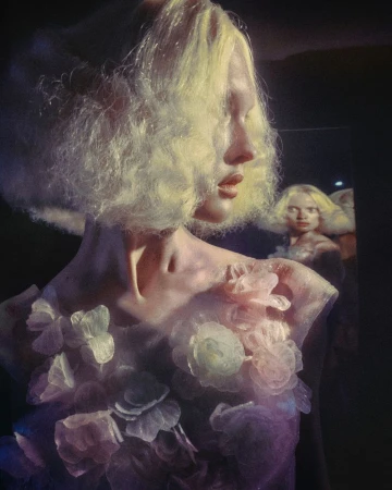 A woman with blonde hair and floral attire poses indoors with a blurred reflection in the background, showcasing an artistic and ethereal quality.