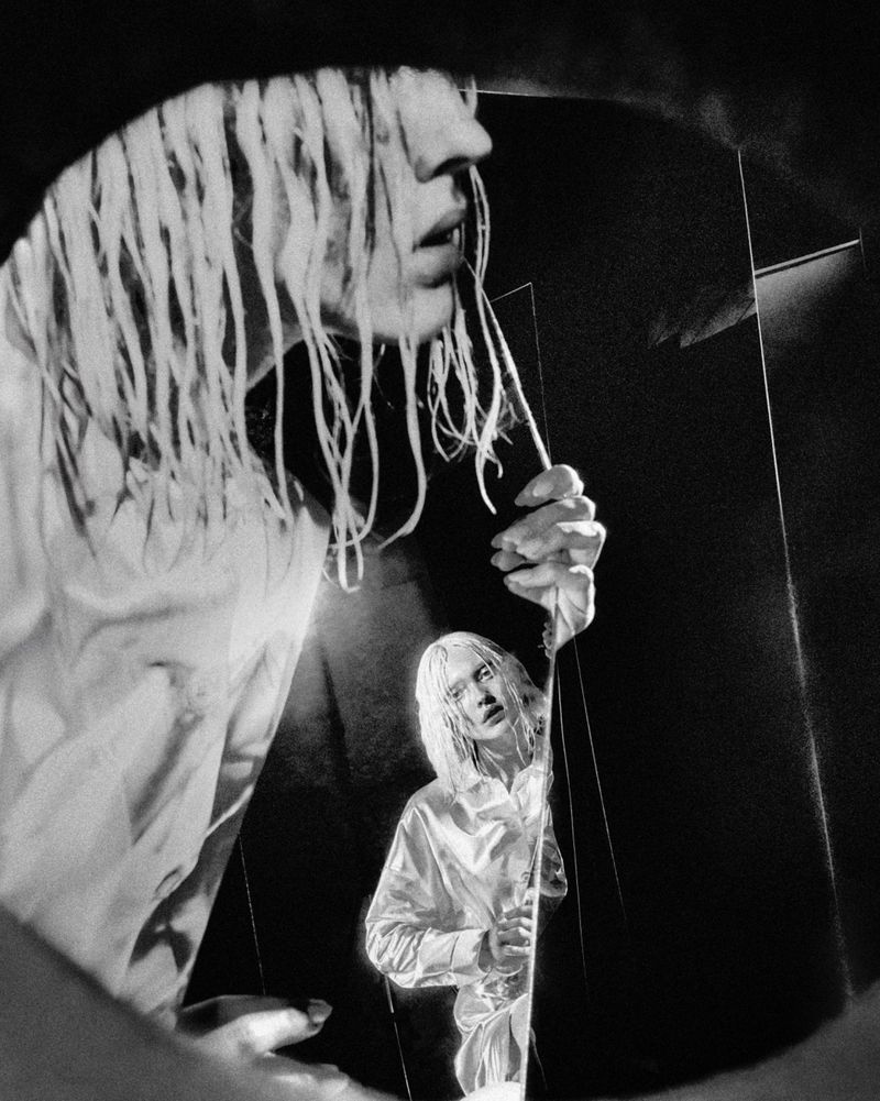Monochromatic fashion shot of a young adult with bleached hair holding a mirror reflecting their face, wearing a white top indoors.