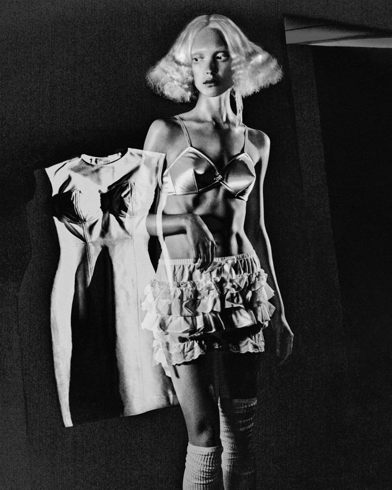 Monochrome portrait of a slender woman in lingerie, knee socks, and an unusual hairstyle, next to a vintage dress on display.