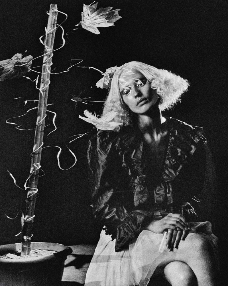 Black and white portrait of a woman with light hair wearing a ruffled shirt and light skirt, seated next to a plant.