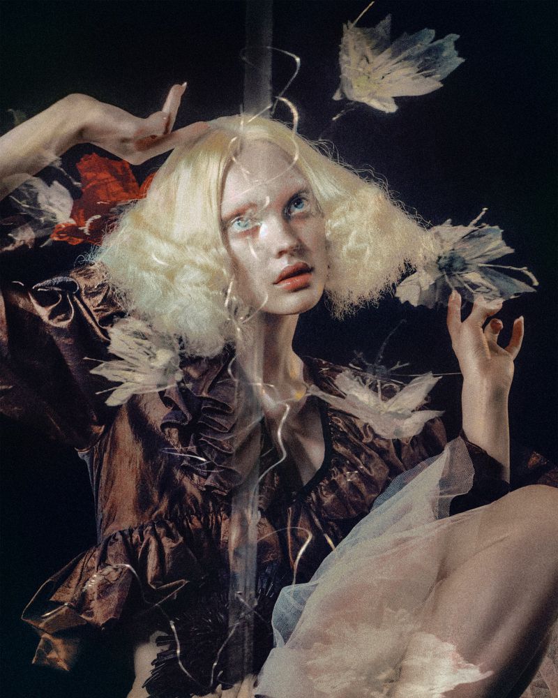 Woman with pale skin poses with dramatic makeup and a voluminous blond wig while wearing a ruffle-necked dress with sheer sleeves.