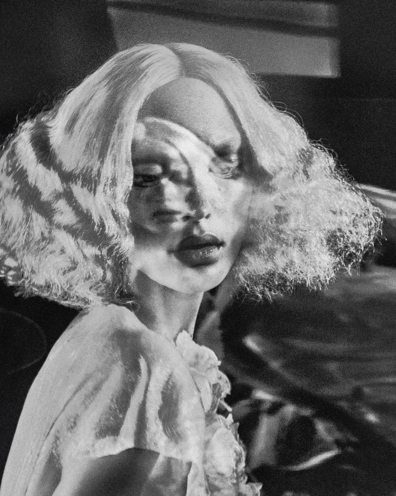Artistic portrait of a woman with blonde curly hair and a frilly white top, set against a dark and blurry backdrop in monochrome.