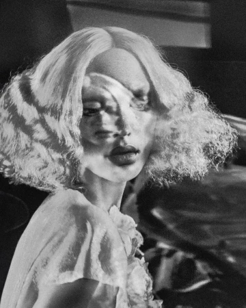 Artistic portrait of a woman with blonde curly hair and a frilly white top, set against a dark and blurry backdrop in monochrome.