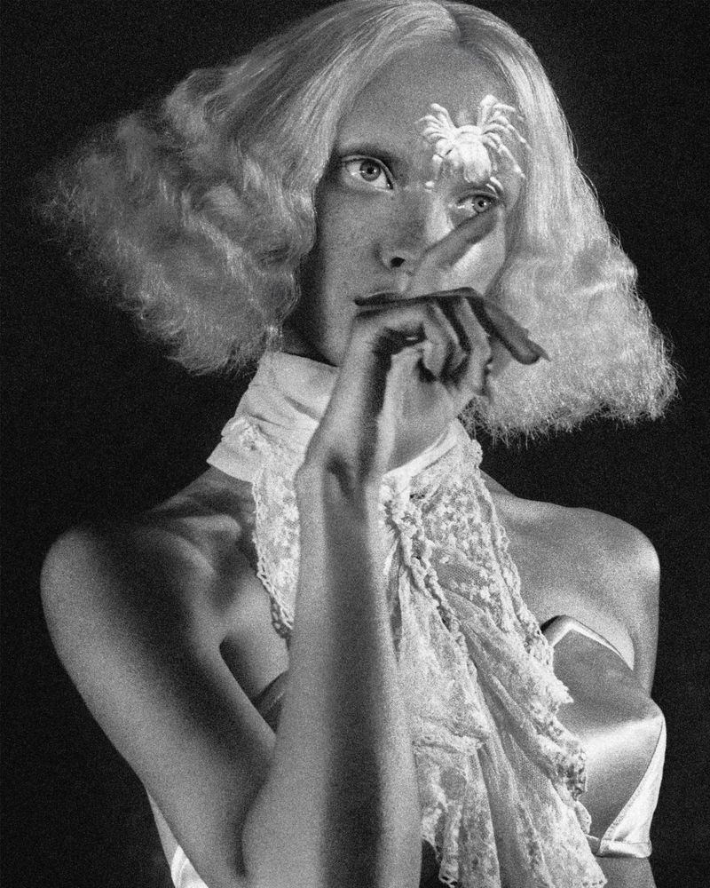 Monochrome portrait of a young woman with wavy blonde hair and a lace scarf, featuring a spider embellishment on her forehead.