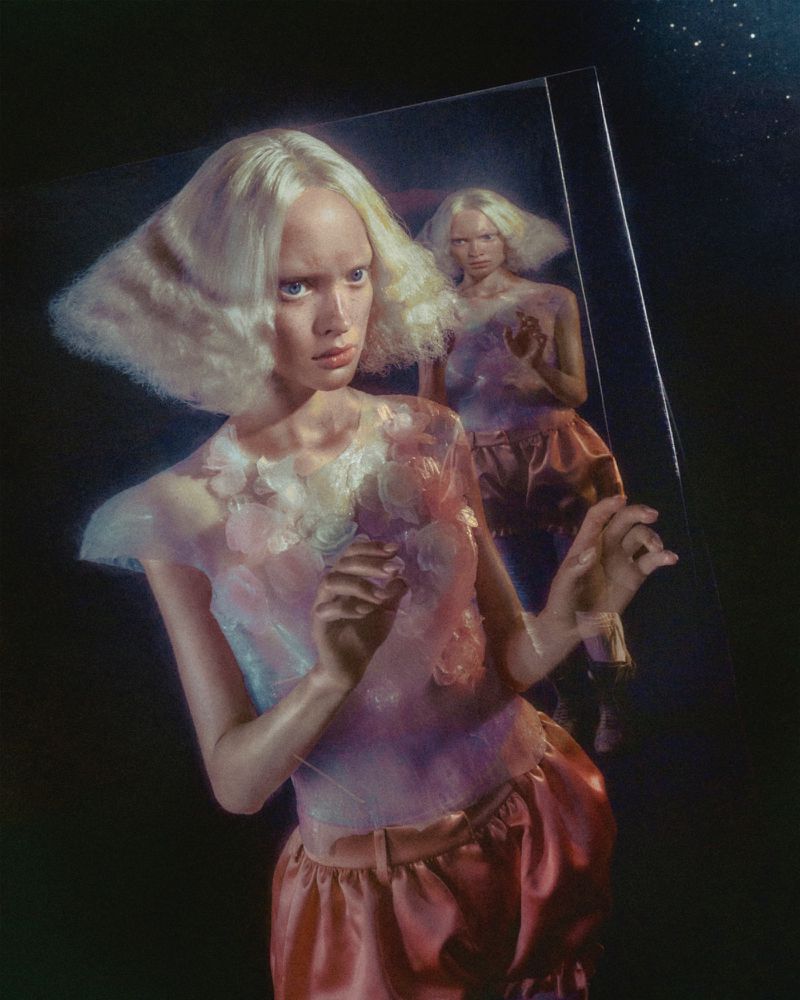 A young woman with bleached blond hair and pale complexion stands in front of a mirror reflecting her image in a fashion editorial.