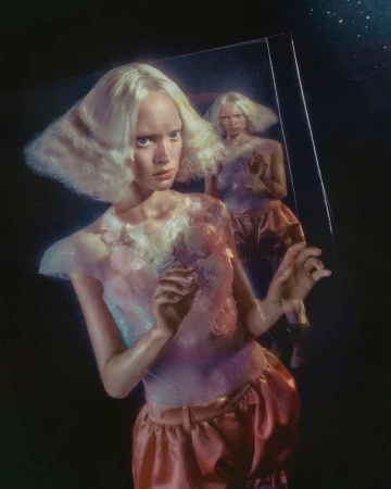 A young woman with bleached blond hair and pale complexion stands in front of a mirror reflecting her image in a fashion editorial.