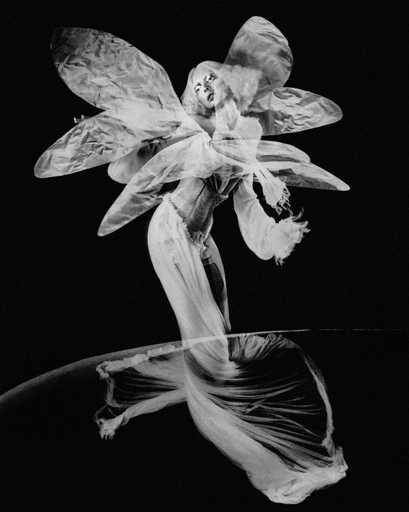 Artistic black and white portrait of a woman in a dress with flower-like embellishments, posing against a stark black background.