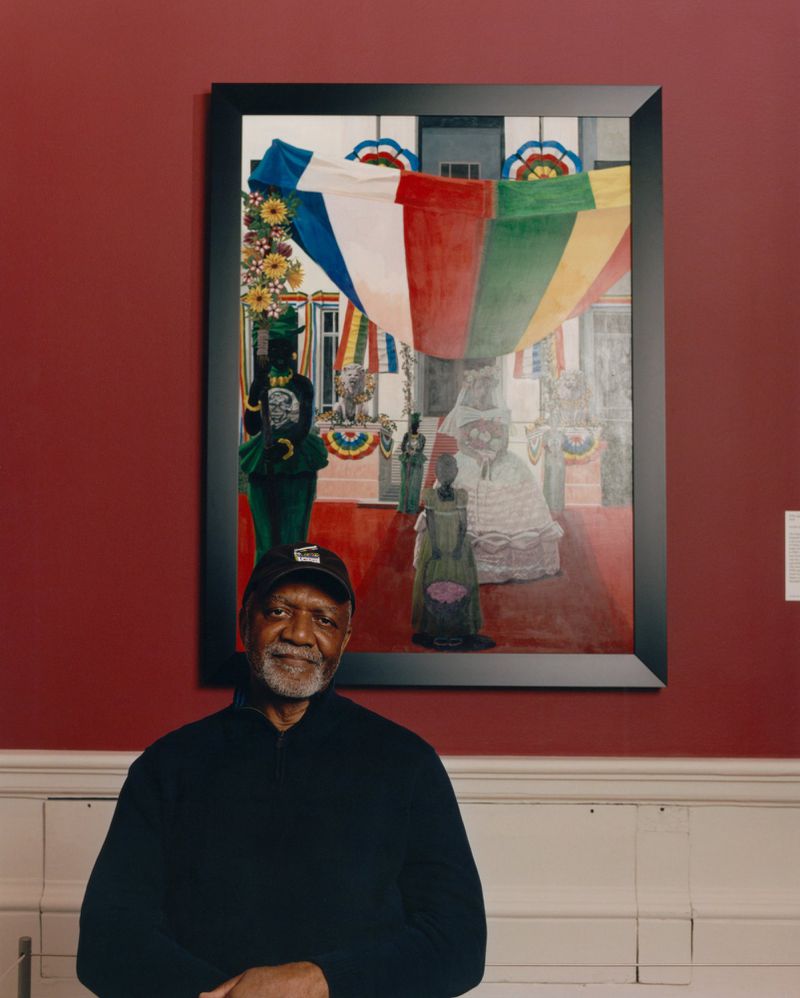 A man in a black cap stands in front of a colorful painting of figures and flags in an indoor setting with red and white walls.