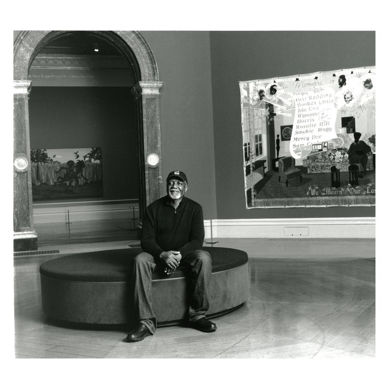 Adult man sits on bench in museum gallery; black and white photograph of man in jeans, sweater and baseball cap, art in background.