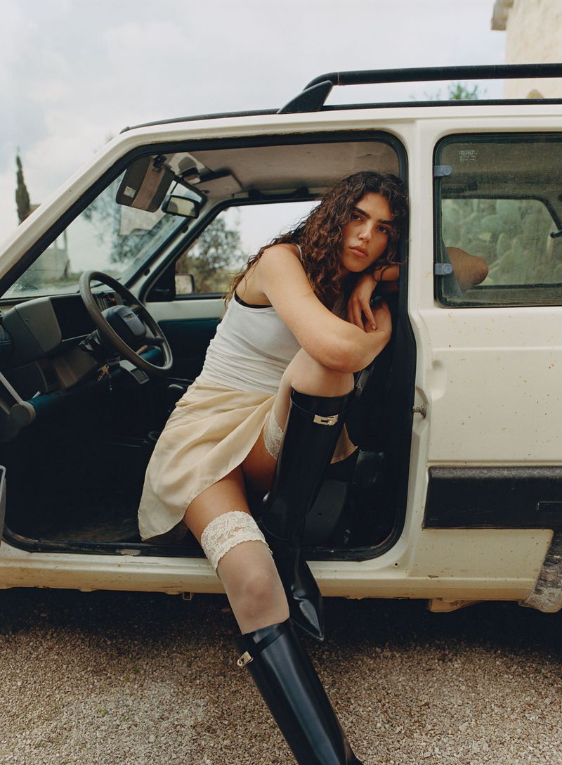 A woman with curly hair sitting in an open car door, wearing black boots, a short skirt, and a tank top in a rural setting.