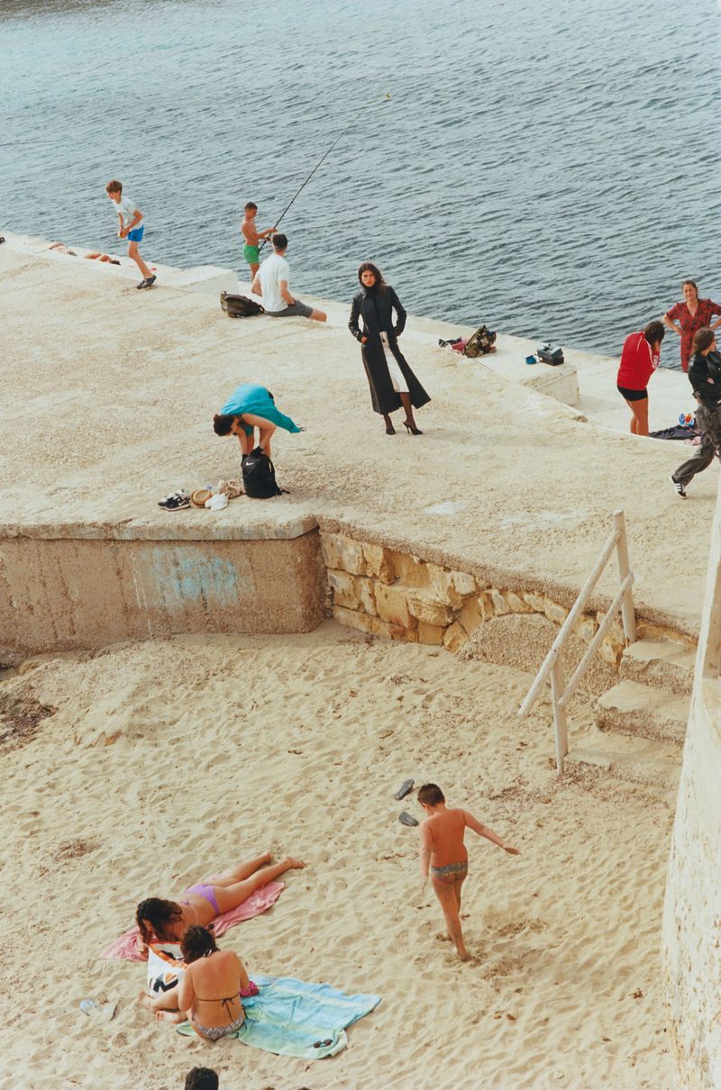 People relax by the ocean on a sunny day: a woman in a black coat, sunbathers, and children playing on a sandy beach by a concrete pier.