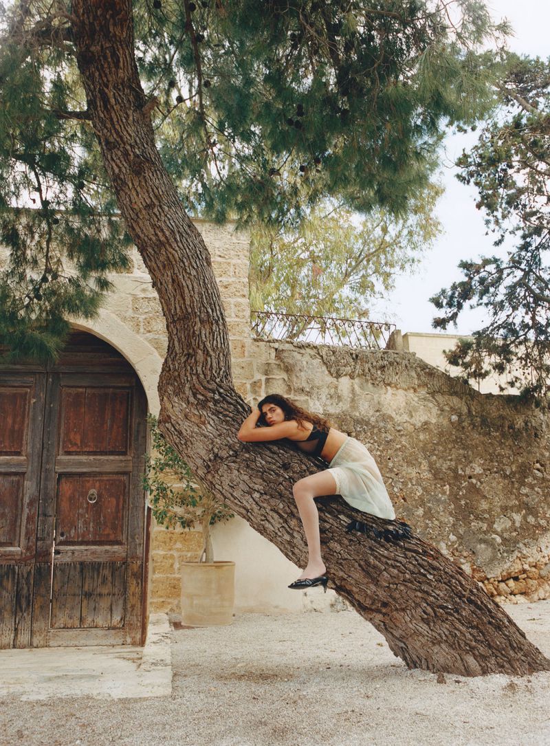 Adult female posing on a tree trunk in a courtyard with a stone building and wooden door in the background, styled with a bra top and skirt.