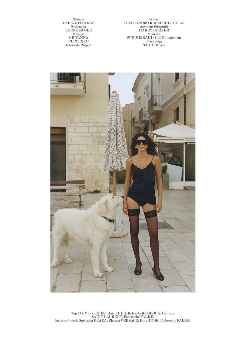 Model in lingerie poses with a Great Pyrenees dog in an outdoor setting with building facades and an umbrella in the background.