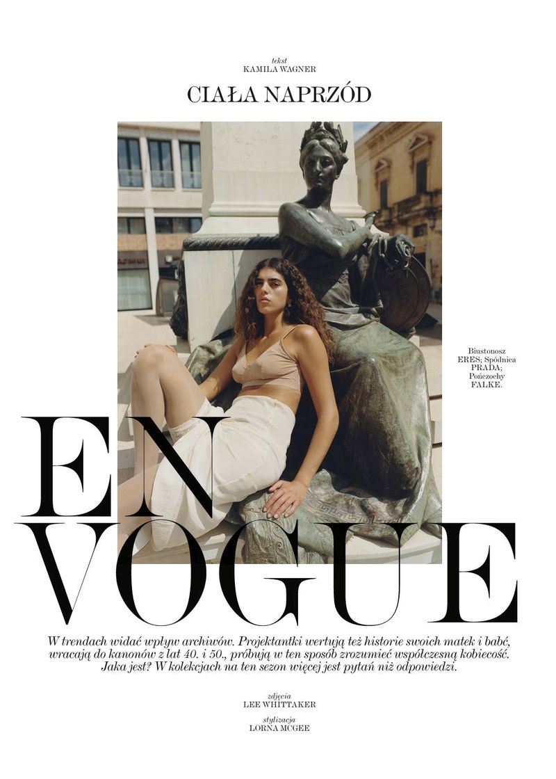Woman poses next to outdoor statue for Vogue magazine cover. Image includes neutral tank top, skirt, city building, and brand logos.
