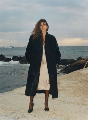 Model Sun Mizrahi wearing a long black coat and champagne slip dress standing along a rocky coast overlooking the ocean.