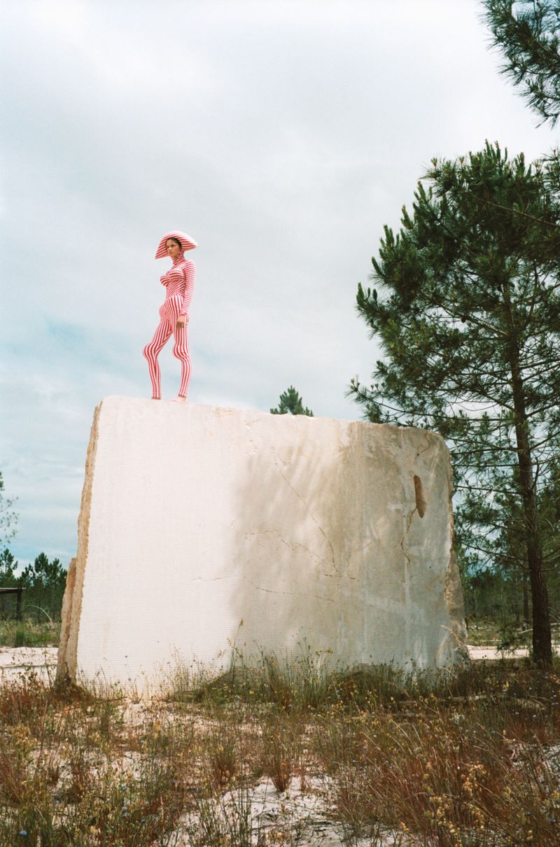Woman in pink striped bodysuit and hat posing atop concrete structure with trees and grassy landscape, outdoors on cloudy day.