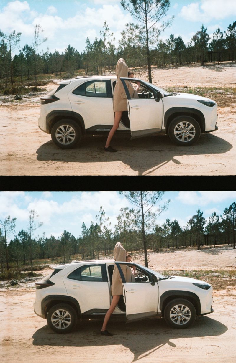 White SUV parked on sandy terrain with a woman opening the driver's side door, backed by a forest under a blue sky.