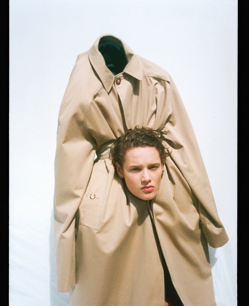 Adult male with short, wavy hair poses wearing a tan trench coat against a white background in a fashion photography studio setting.