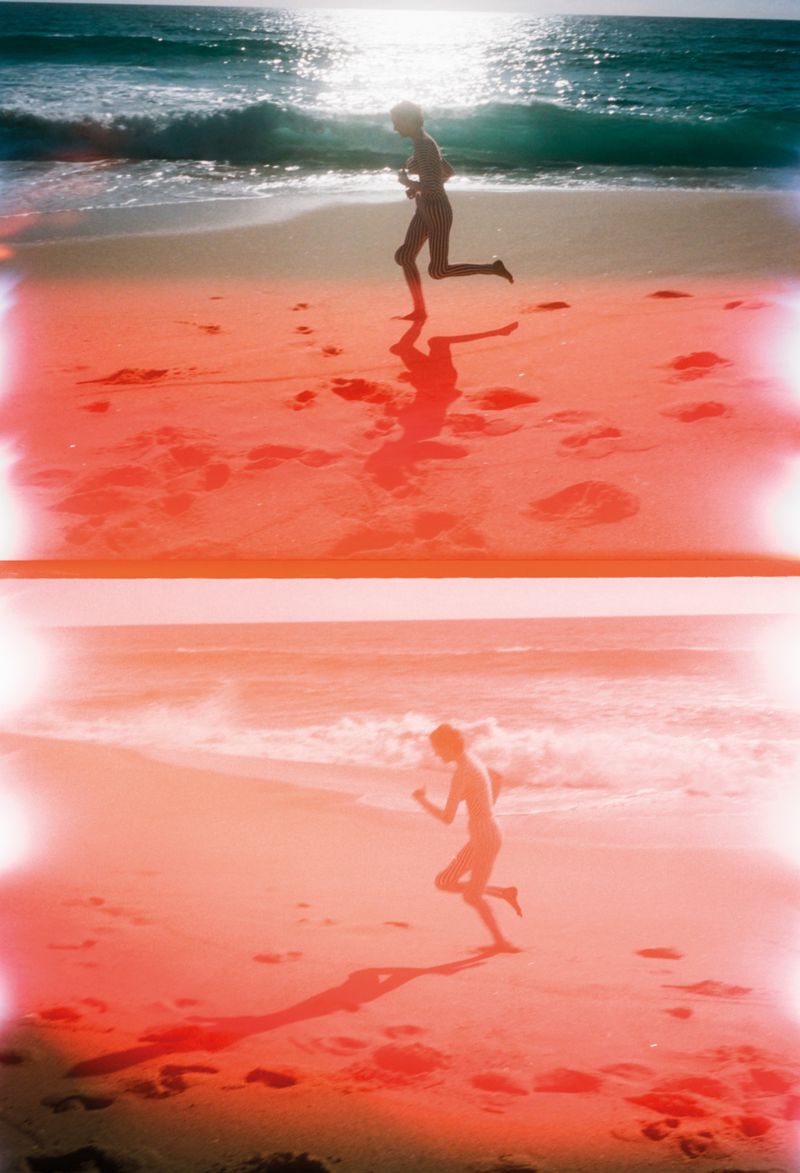 A woman runs on a sunny beach next to the ocean with waves, depicted in a film still style with red and white light leaks.