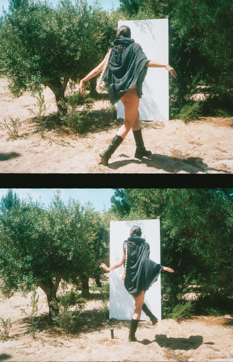 Fashion shot in an outdoor setting with a person wearing a grey poncho, boots, and striking a pose against a white panel.
