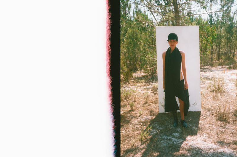 Woman posing in a black dress and hat in a rural outdoor setting. She stands in front of a white board with sparse trees behind.