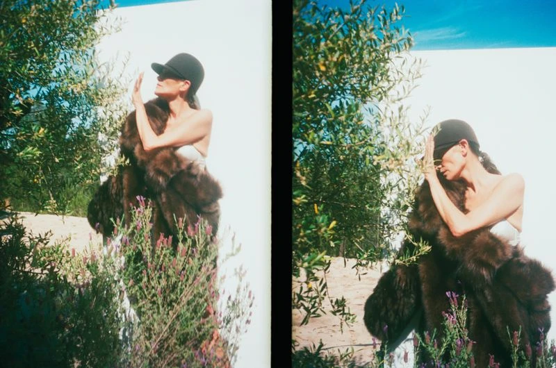 A woman in a fur coat and baseball cap stands outdoors among greenery against a light blue sky in a fashion editorial style.