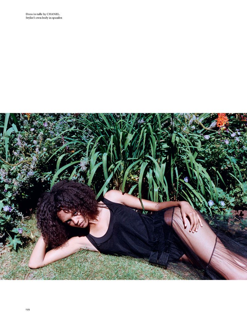 Woman in black dress reclines on grass surrounded by green plants in a garden setting, capturing a serene and elegant fashion moment.