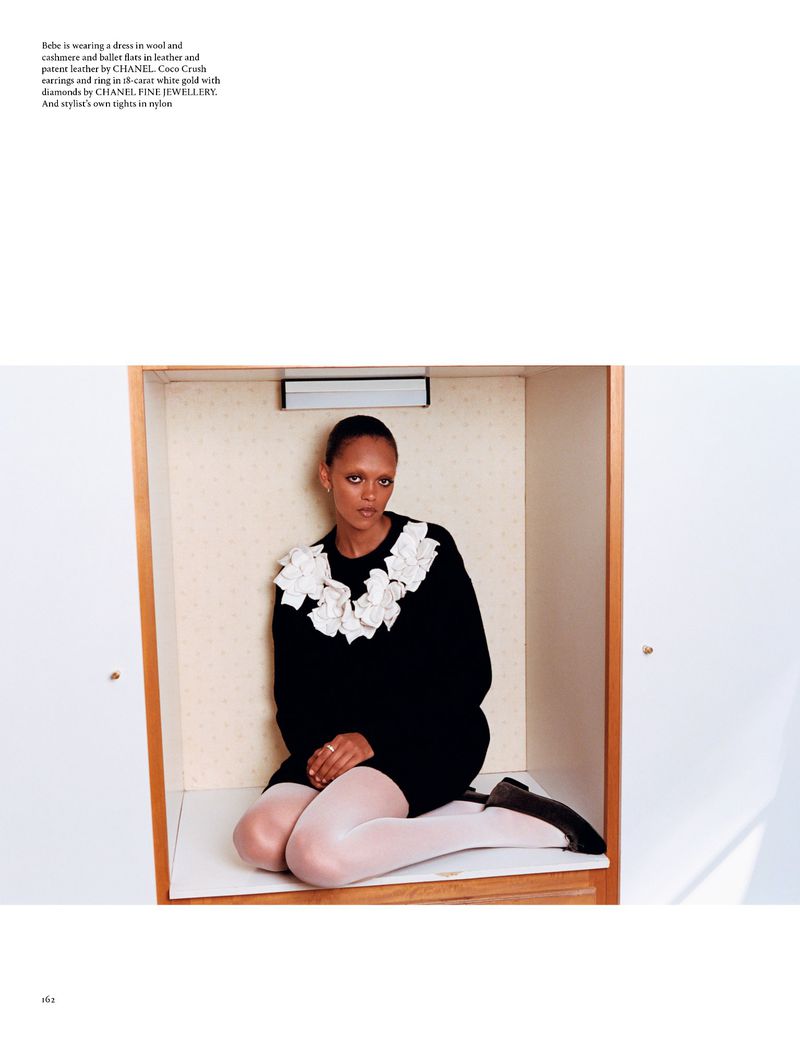Adult female fashion model in a black top with white floral collar detail poses inside of a light beige coloured wardrobe setting.