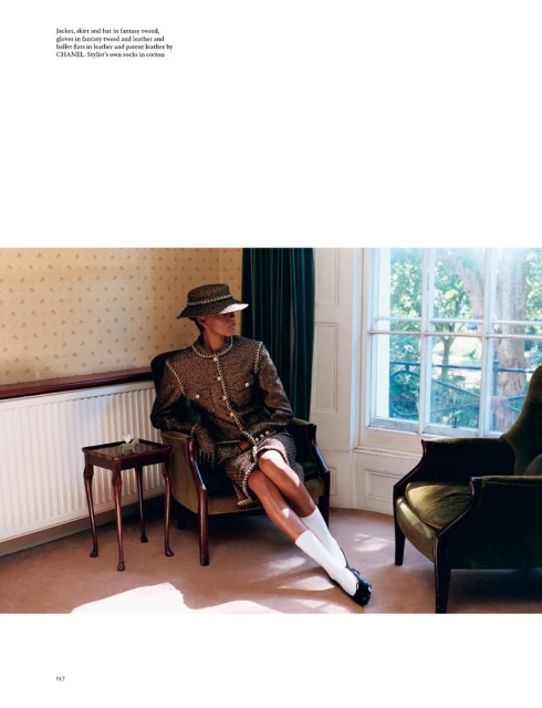 A woman wearing a tweed suit and hat sits in an armchair near a window in an interior space with an additional armchair and side table.