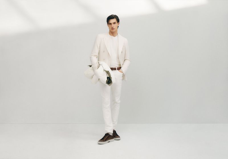 A stylish adult male wearing a cream-colored suit and casual shoes is posing in a studio environment with a light background and shadow.