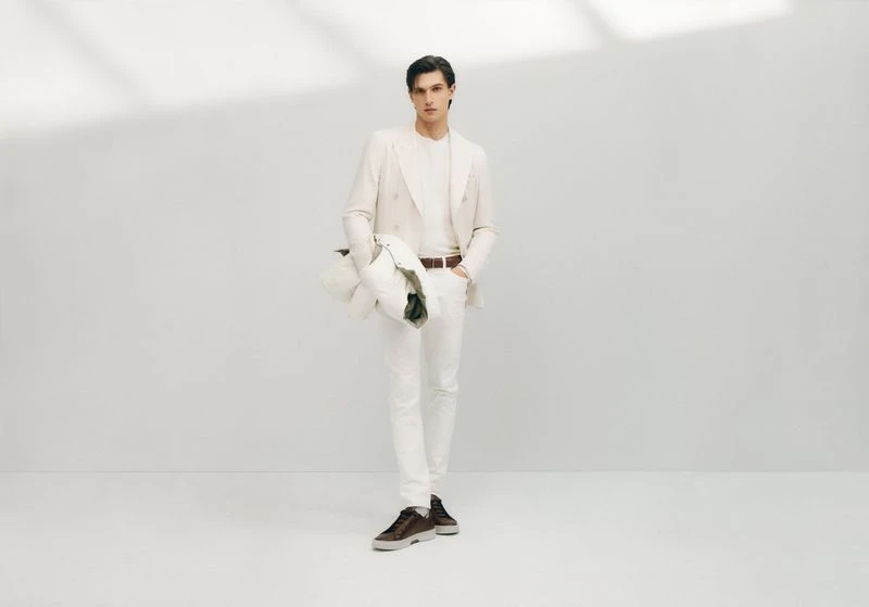 A stylish adult male wearing a cream-colored suit and casual shoes is posing in a studio environment with a light background and shadow.