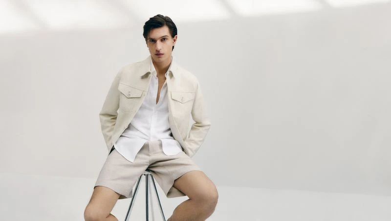 A fair-skinned adult male poses indoors, wearing a white button-down shirt, jacket, and beige shorts while sitting on a black stool.