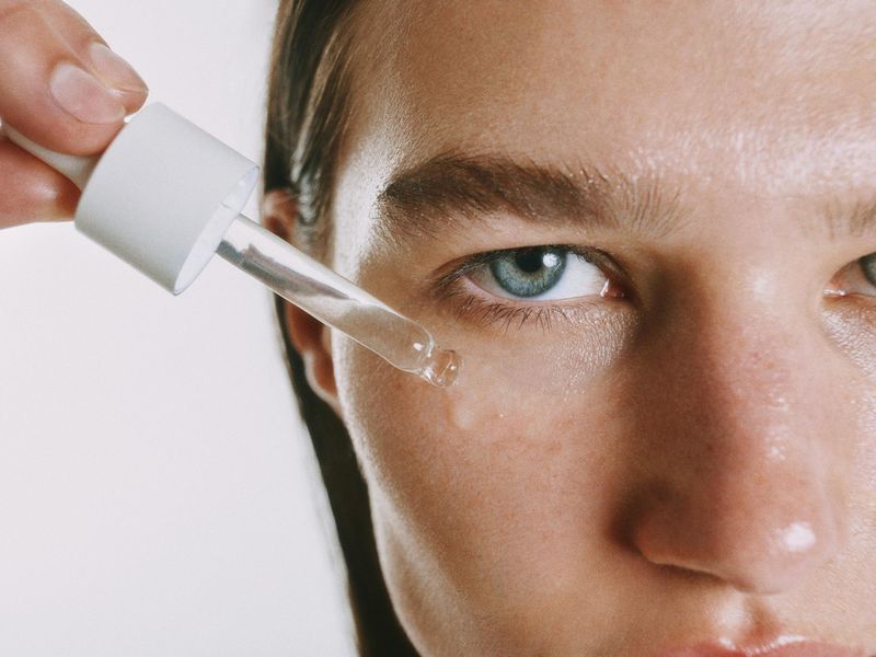 Close-up of a person applying liquid skincare serum to their cheek with a glass dropper against a white backdrop.