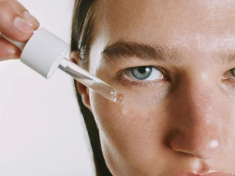 Close-up of a person applying liquid skincare serum to their cheek with a glass dropper against a white backdrop.