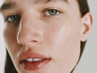 Extreme close-up of a person's face showing dewy, hydrated skin, glossy lips, and green eyes.