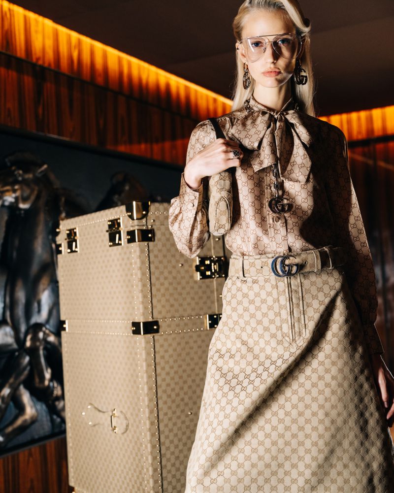 Fashionable blonde woman in patterned blouse and skirt with large beige luggage and vintage decor in indoor setting, accessorized with jewelry.