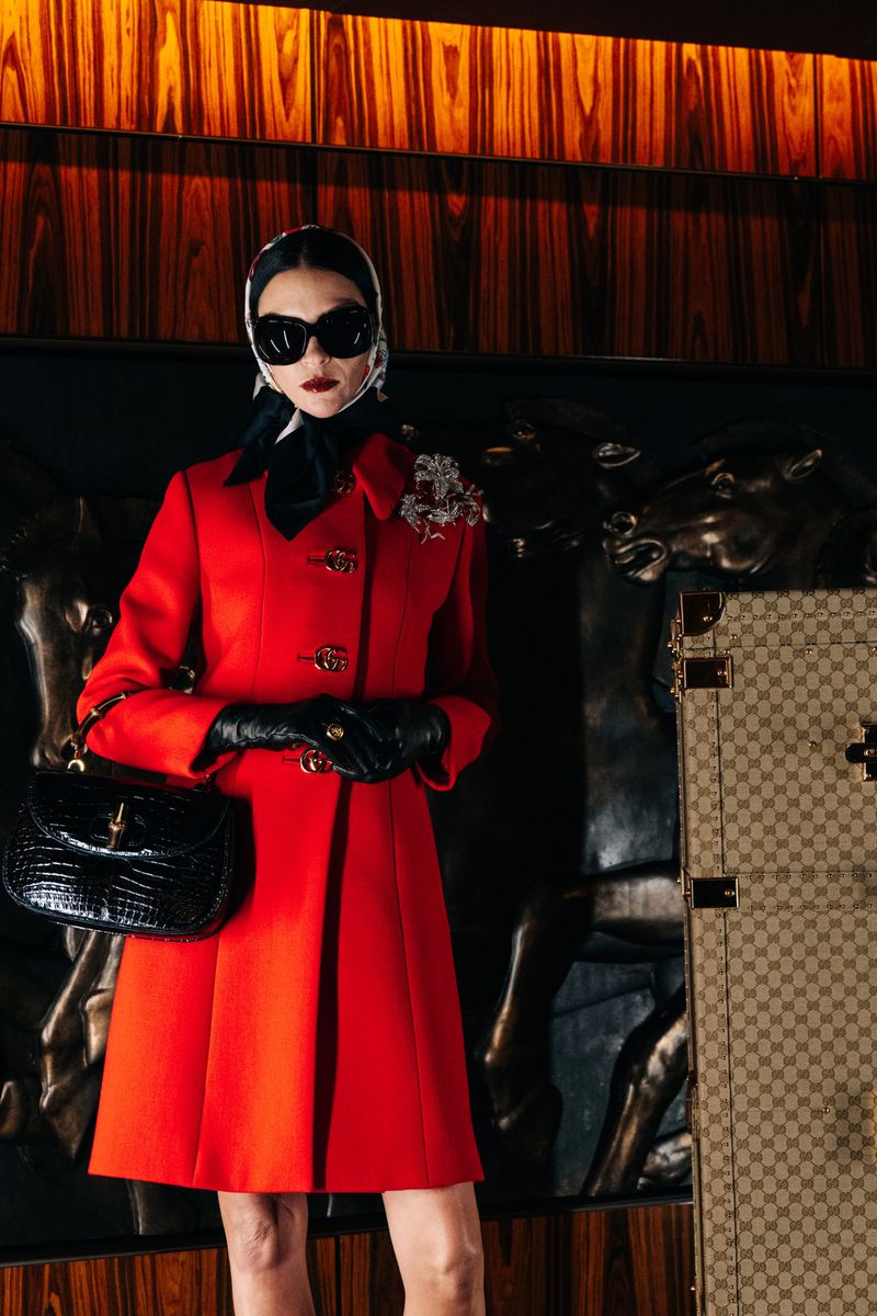 Fashion shot featuring an adult female in red coat, scarf, sunglasses, gloves, carrying a black purse, standing by luggage.