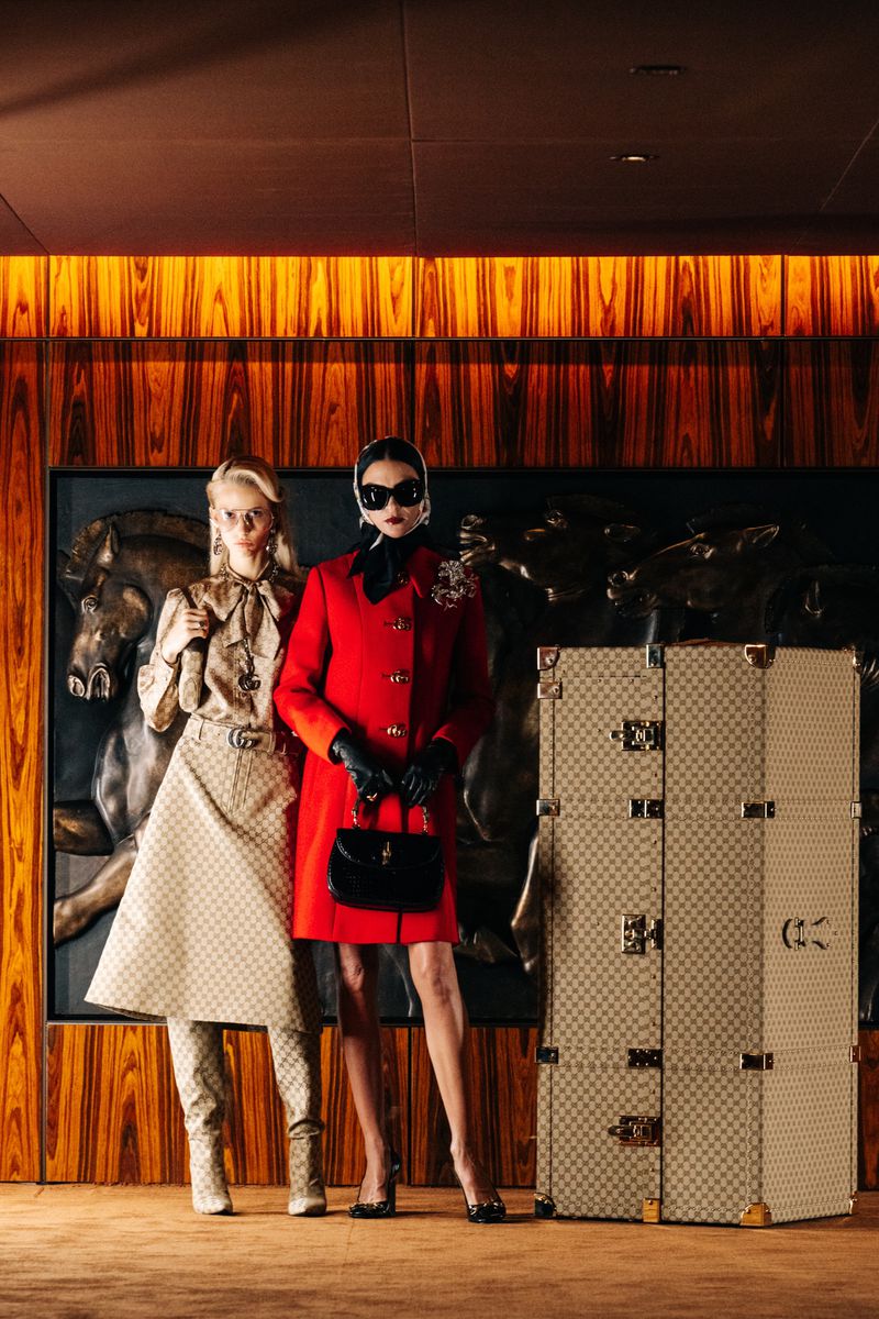 Two women in vintage-inspired outfits pose next to luggage with a horse art background, showcasing a blend of classic fashion and artistic flair.