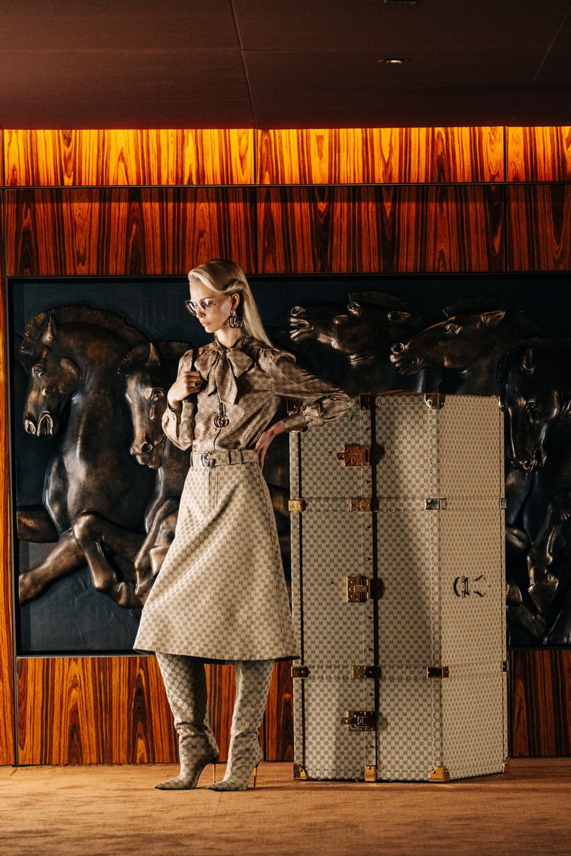 A woman in patterned clothing stands next to a stackable suitcase in a luxurious interior with horse art on the wall and warm lighting.