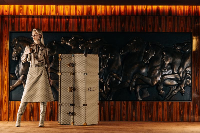 A fair-skinned model poses in a patterned skirt and blouse, next to a vintage-style trunk against a backdrop of wood paneling and horse art.