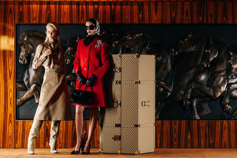 Two women in stylish coats pose beside a Gucci trunk in a vintage setting, showcasing high-fashion apparel and luxury travel accessories.