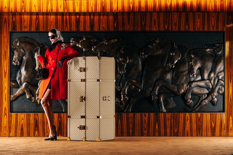 Fashionable woman poses in a red coat with a vintage trunk. She's wearing sunglasses and a scarf against an elegant wood panel backdrop.
