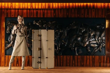 Model in patterned blouse, matching midi skirt, and tall boots standing next to a large standing wardrobe trunk against a wood-paneled wall.