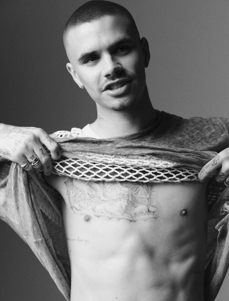 Black-and-white portrait of a shirtless man with tattoos, short hair, and a neutral expression, standing indoors against a gray backdrop.