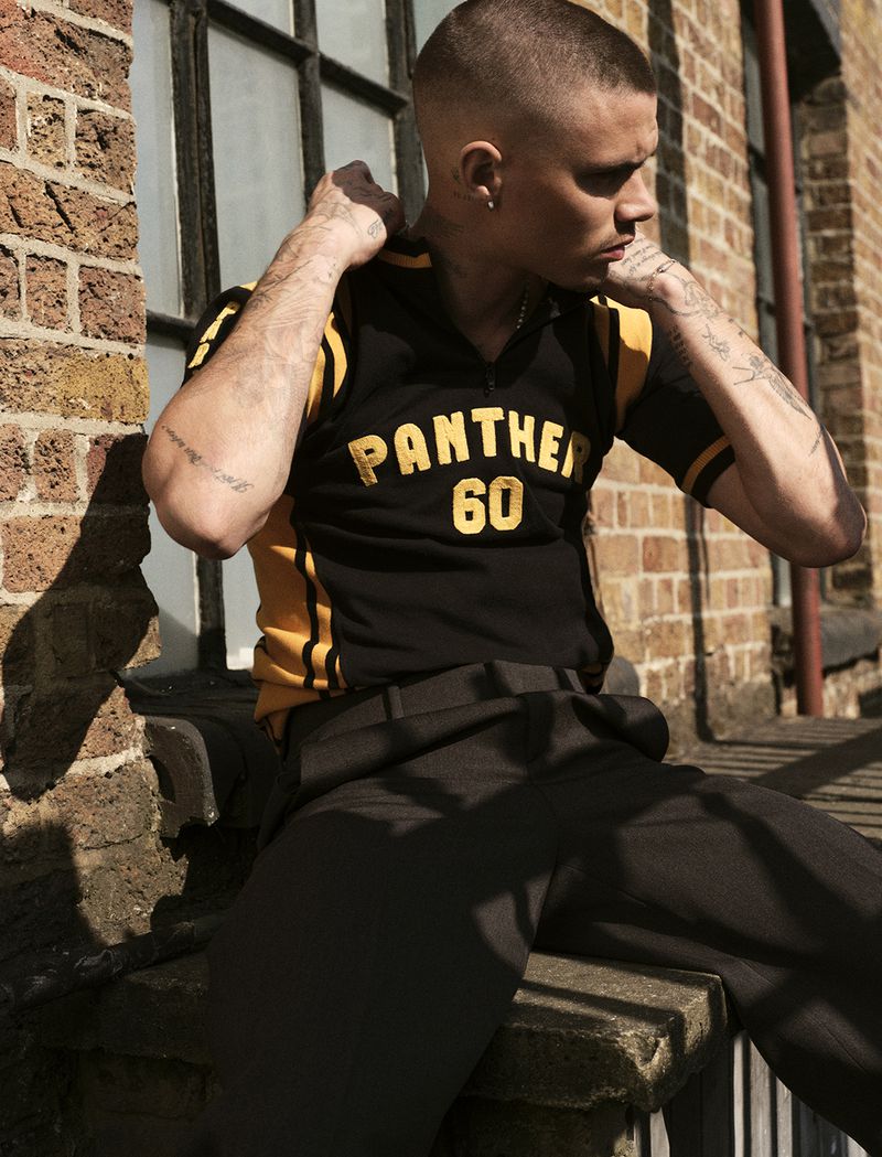 Young adult male posing outdoors in a black and gold jersey, seated against a brick wall. He has short hair and visible tattoos.