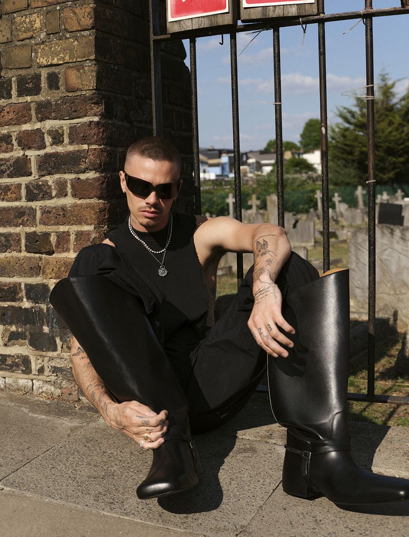 A man with sunglasses is squatting against a brick wall and an iron fence wearing black boots, pants, and a tank top outdoors in daylight.