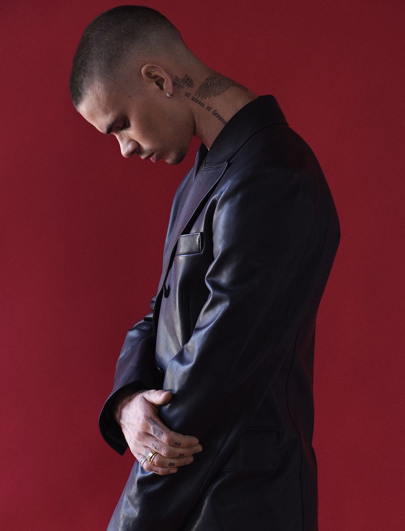 Man in a dark suit standing against a red background with his head down. He has short hair, a ring, and neck tattoo.