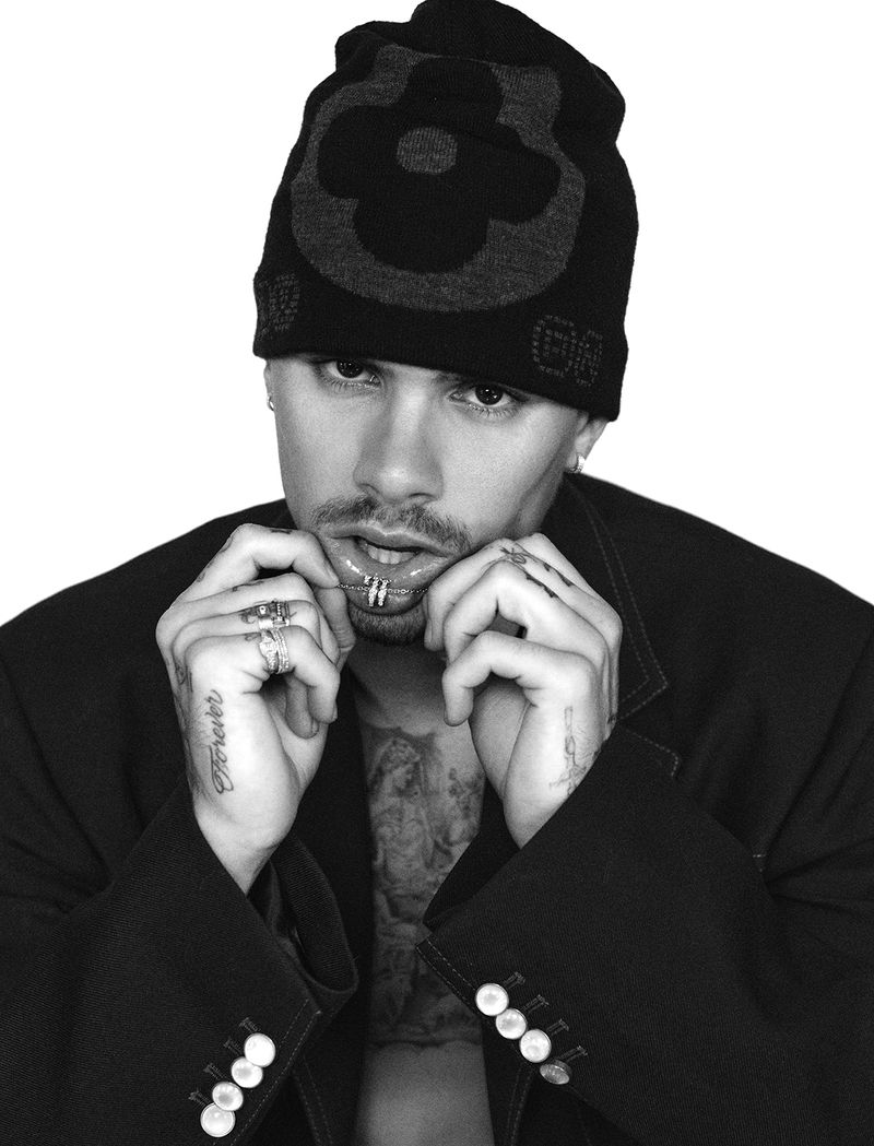 Black and white portrait of a young adult man wearing a black beanie and blazer, with multiple rings and a lip piercing.