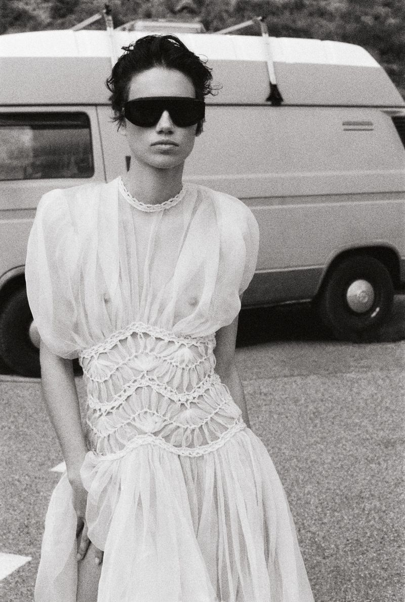 Woman in a white dress and sunglasses stands in front of a van on a road. Black and white fashion shot. She poses in daylight.